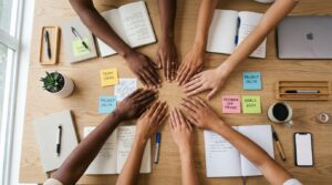 Team members place their hands together on a table surrounded by notebooks and project notes.